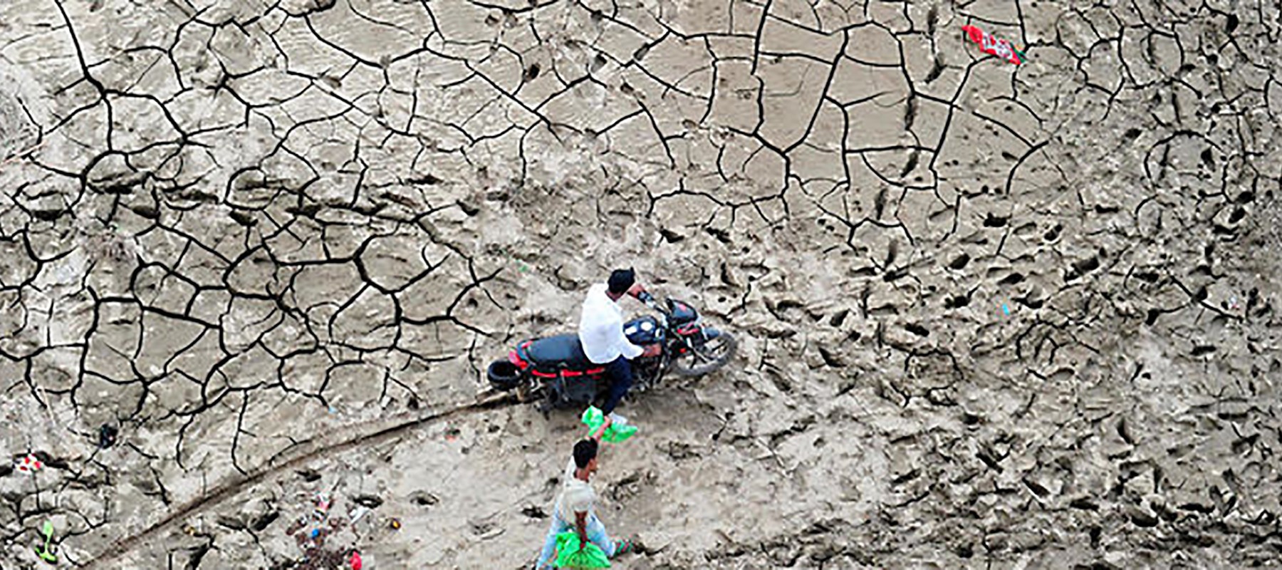 Motorbike and man carrying water cans walking on dry, cracked soil